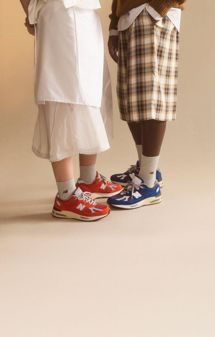 Waist down view of a man and woman wearing New Balance Made in UK sneakers standing in a beige studio environment. 