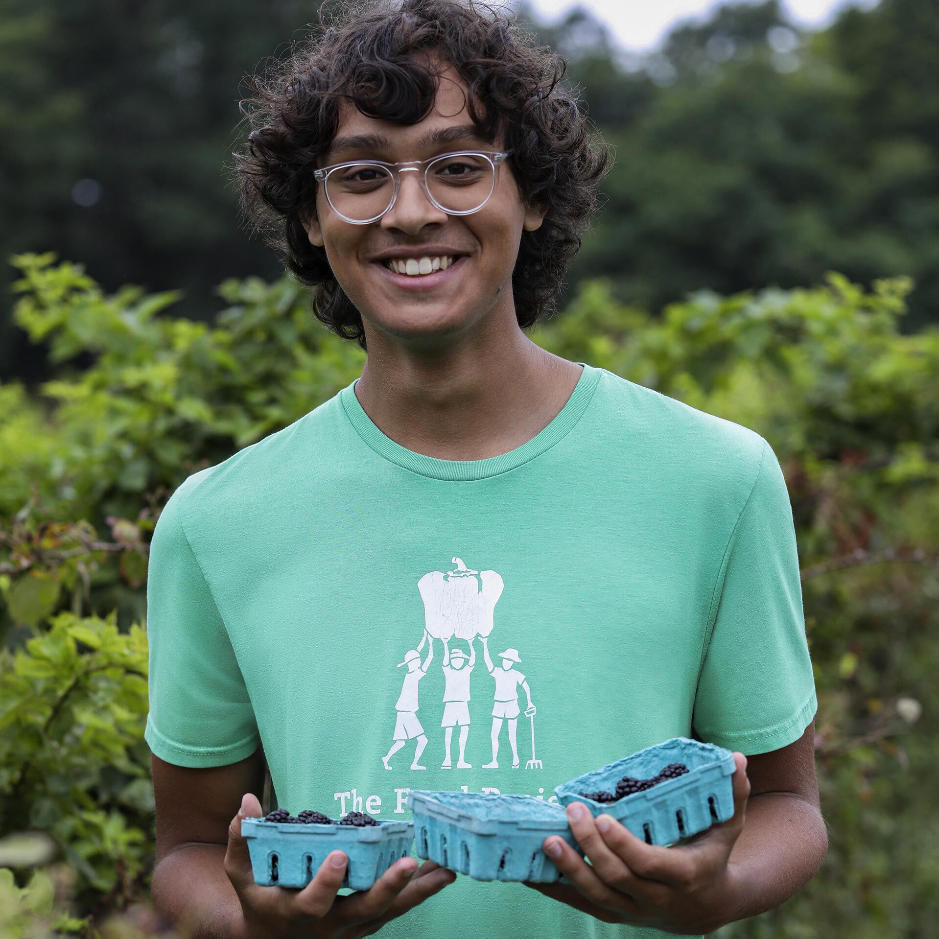 A medium-skinned, male-presenting teen holding several containers of blackberries he&rsquo;s harvested himself as a farm volunteer.