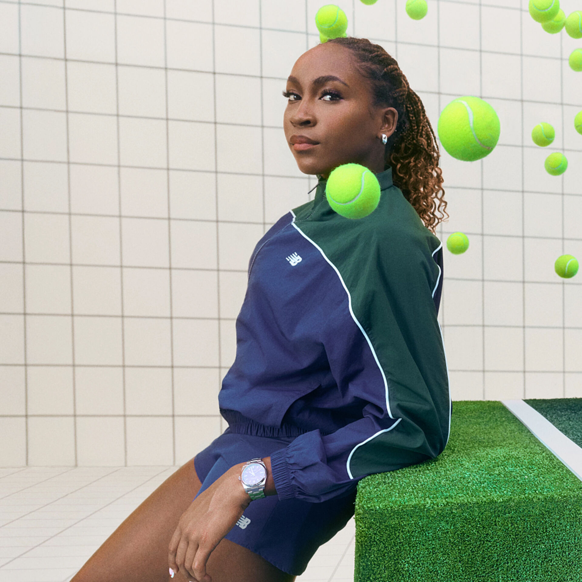 Coco Gauff leaning on a block, surrounded by tennis balls, dressed in her New Balance tennis kit.