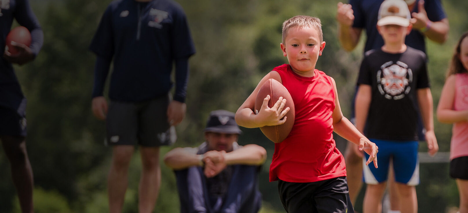A young, light-skinned, male-presenting child running down a field with a football with spectators cheering him on.