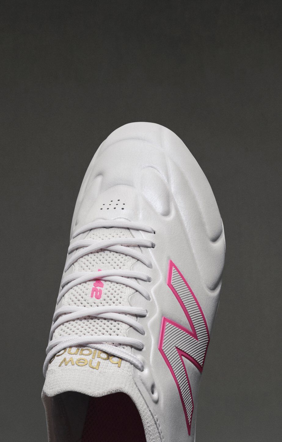 Top‑down close‑up of a white New Balance soccer cleat on a dark gray background, featuring white laces, a textured upper, pink detailing, and a metallic‑outlined pink &ldquo;N&rdquo; logo along the side. 