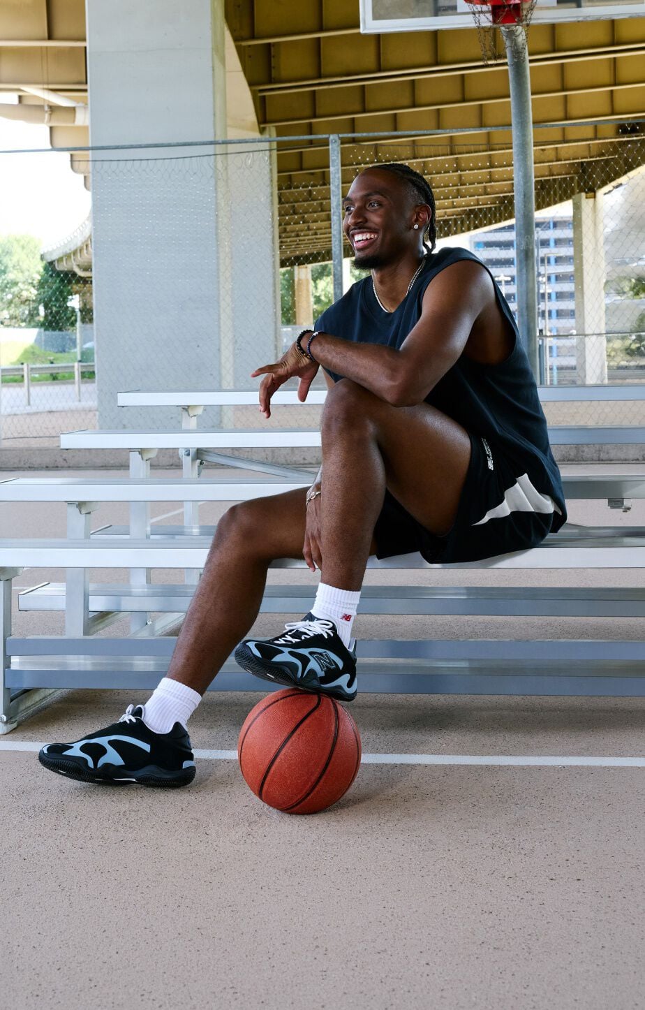 Person seated on outdoor bleachers beneath an overpass with one foot resting on a basketball, showcasing black-and-light-blue athletic sneakers. 