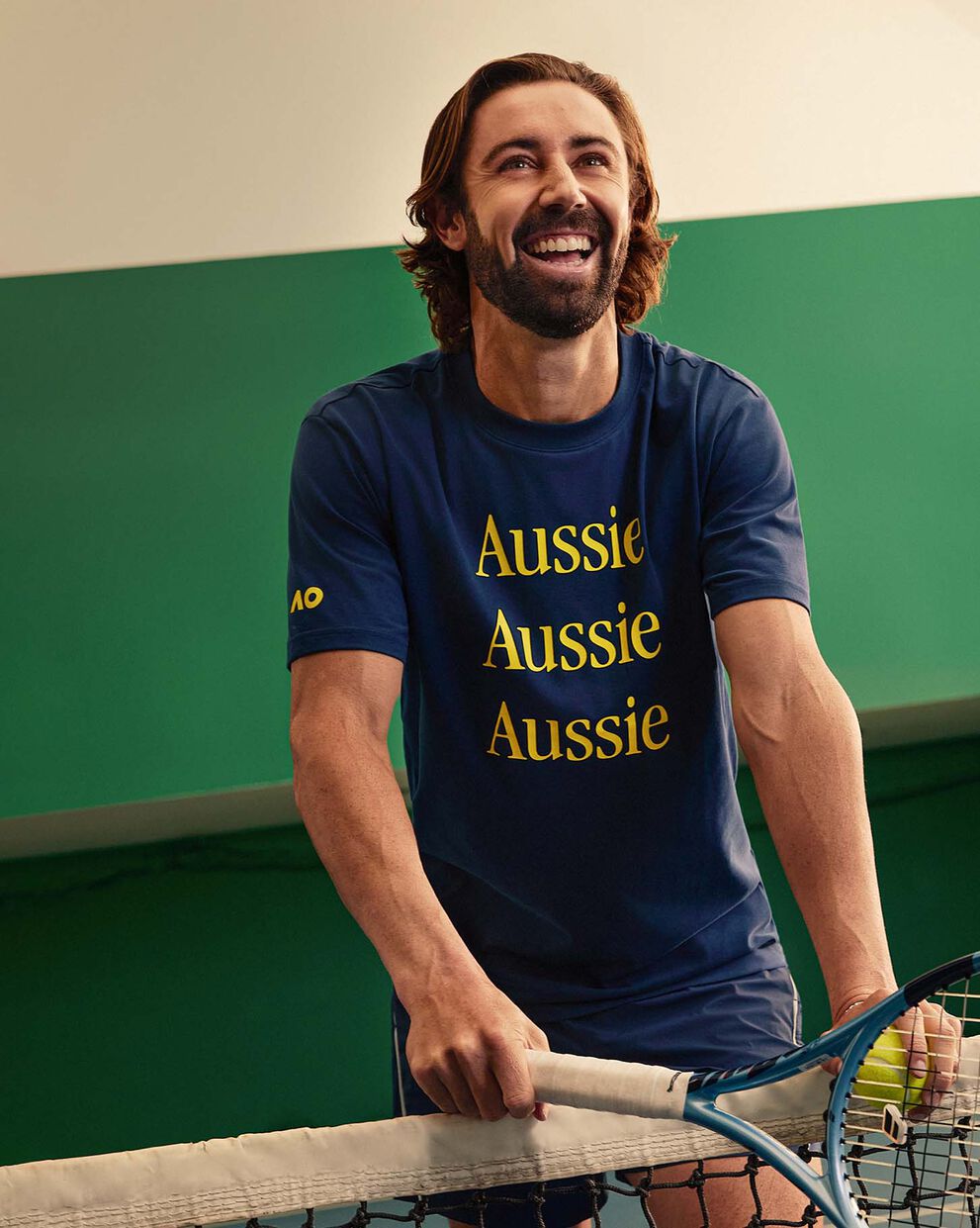 A man smiling at the camera and leaning over a tennis net.