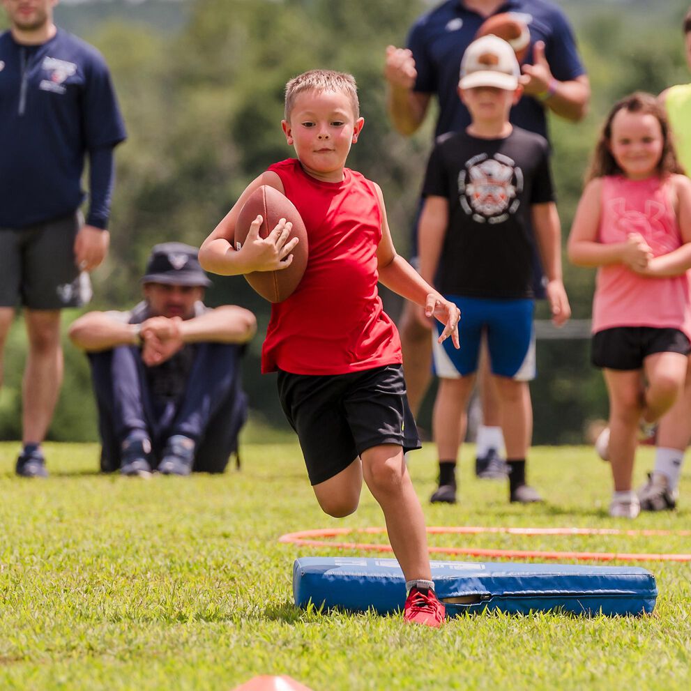 A young, light-skinned, male-presenting child running down a field with a football with spectators cheering him on.