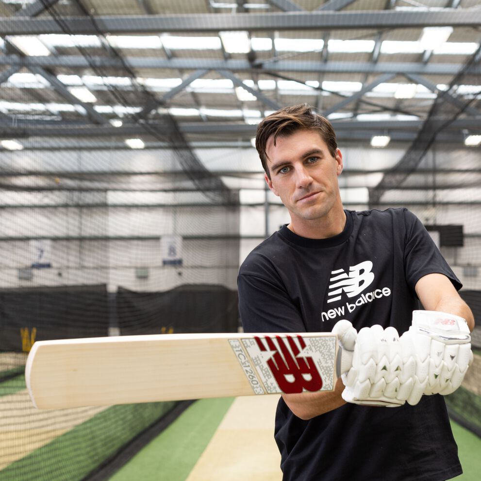 Man standing in indoor cricket complex, swinging New Balance bat, wearing New Balance clothing and cricket gloves