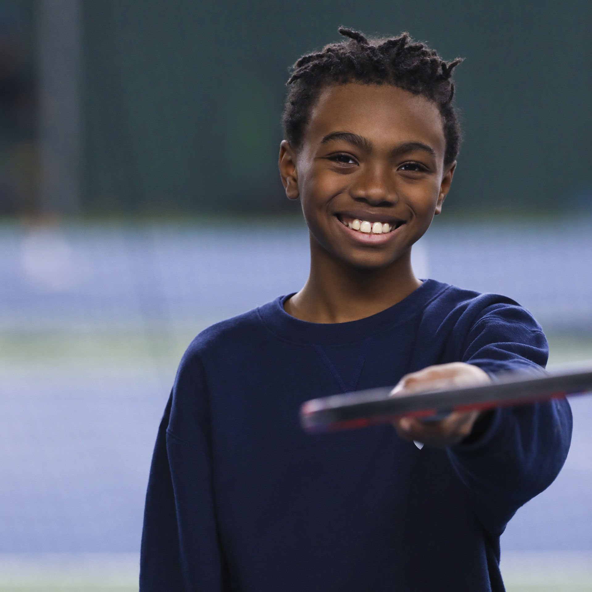 A dark-skinned, male-presenting teen holding a tennis racquet smiling at the camera.