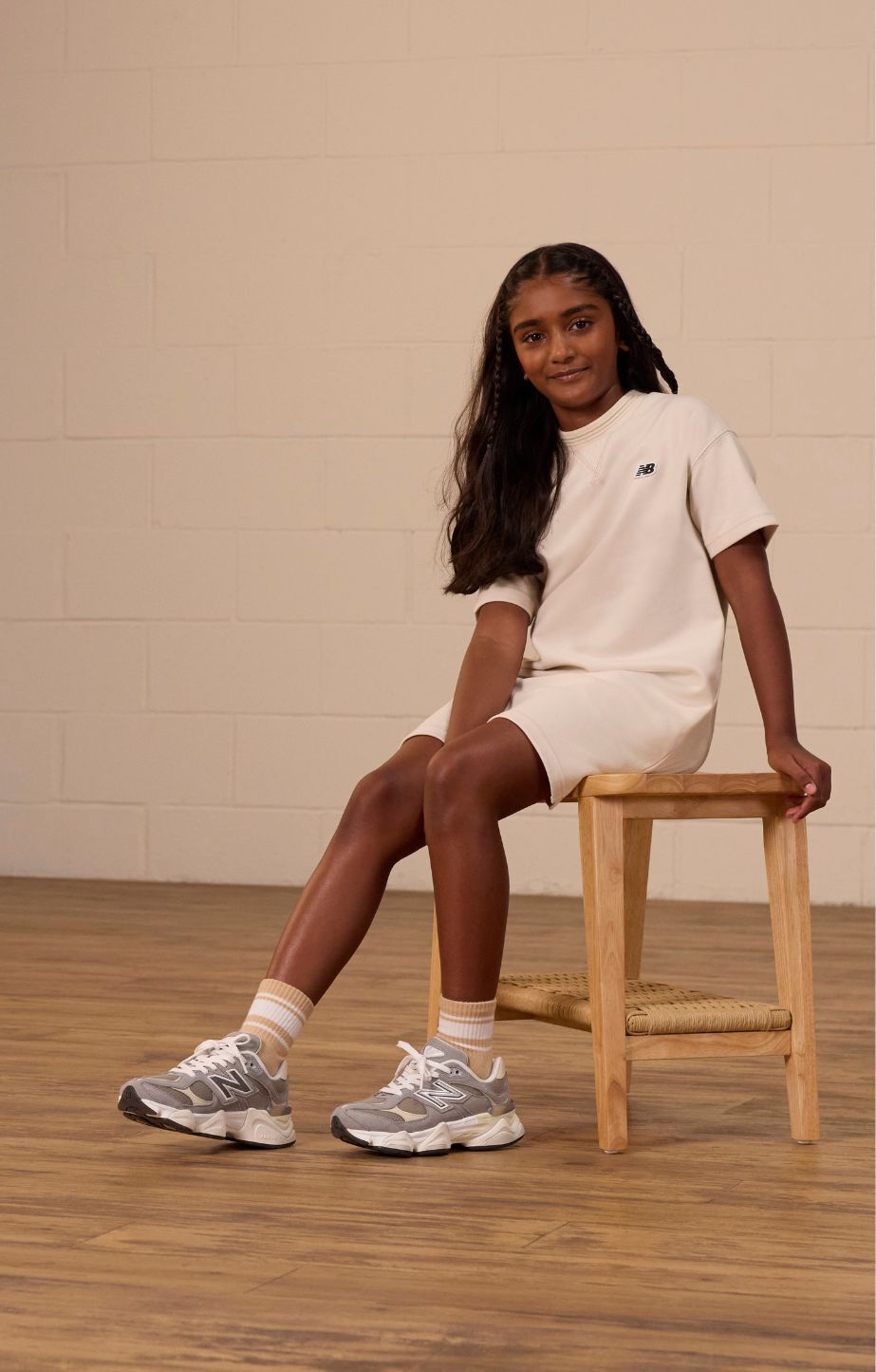 A girl in a light-colored t-shirt dress and grey new balance 9060 sneakers sits on a wooden stool.