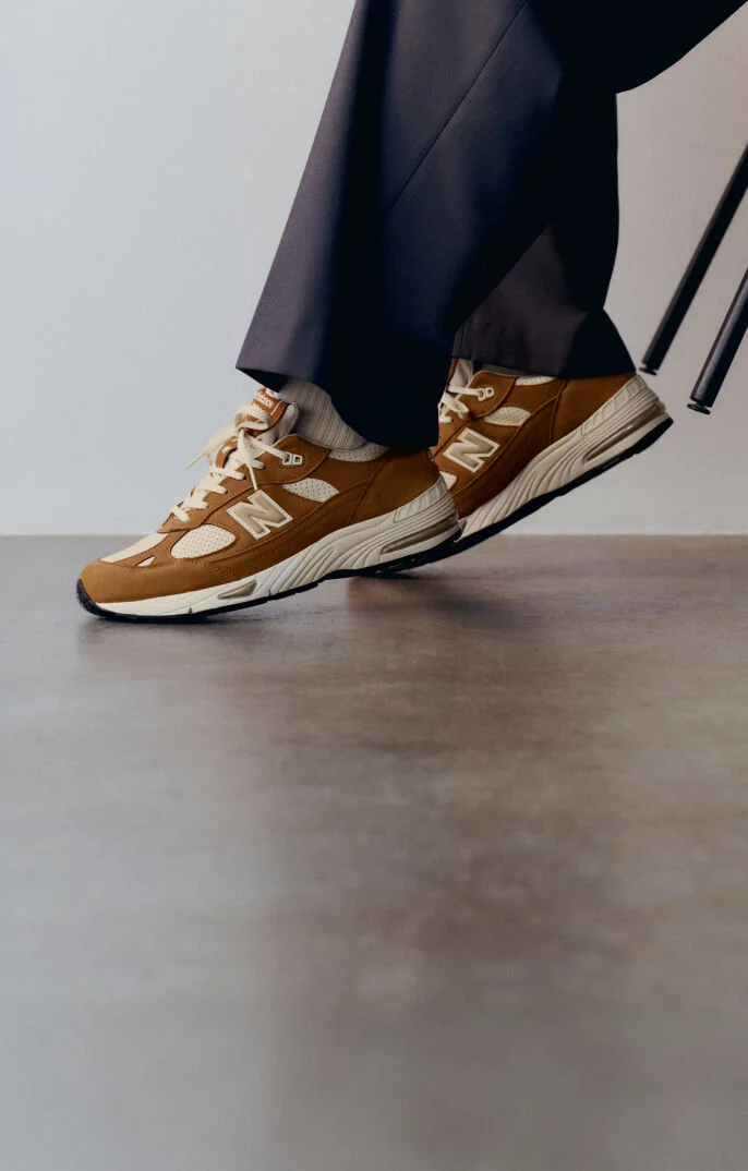 Knee down view of a woman wearing a pair of New Balance Made in UK sneakers in with room with a concrete floor and white walls. 