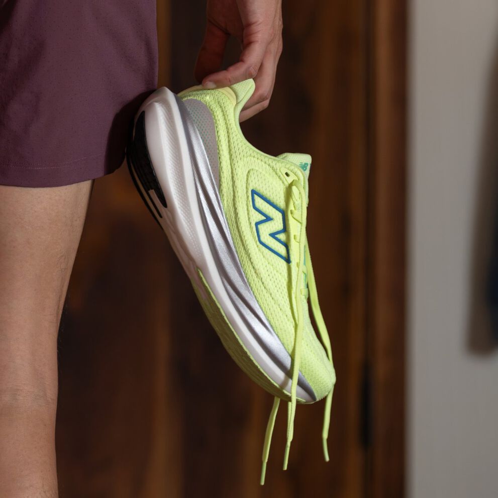 Close-up of a bright yellow-green New Balance running shoe with blue logo and white sole, held by a person wearing maroon shorts. 