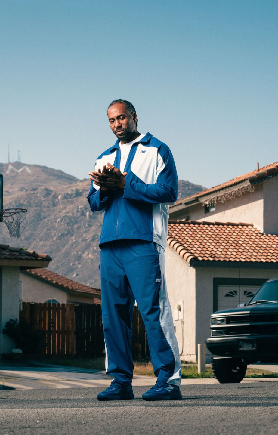 Person wearing a blue and white tracksuit and matching sneakers stands on a suburban street with mountains and houses in the background. 